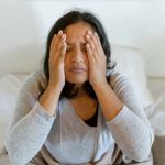 A woman rubs her head as she sits up in bed.
